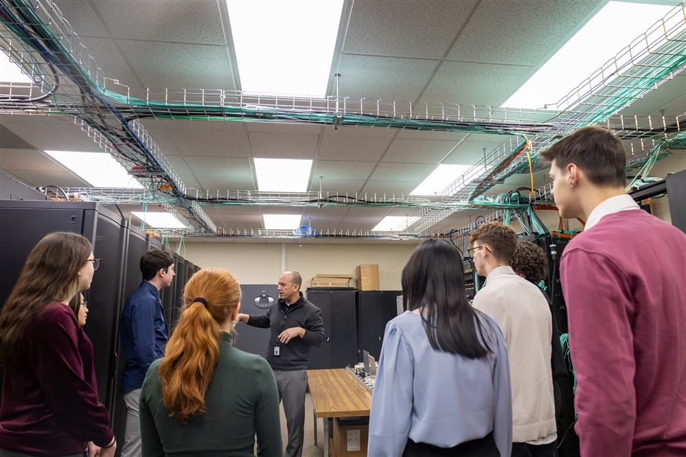 A man is talking to a group of eight students as he shows them the internet server room.